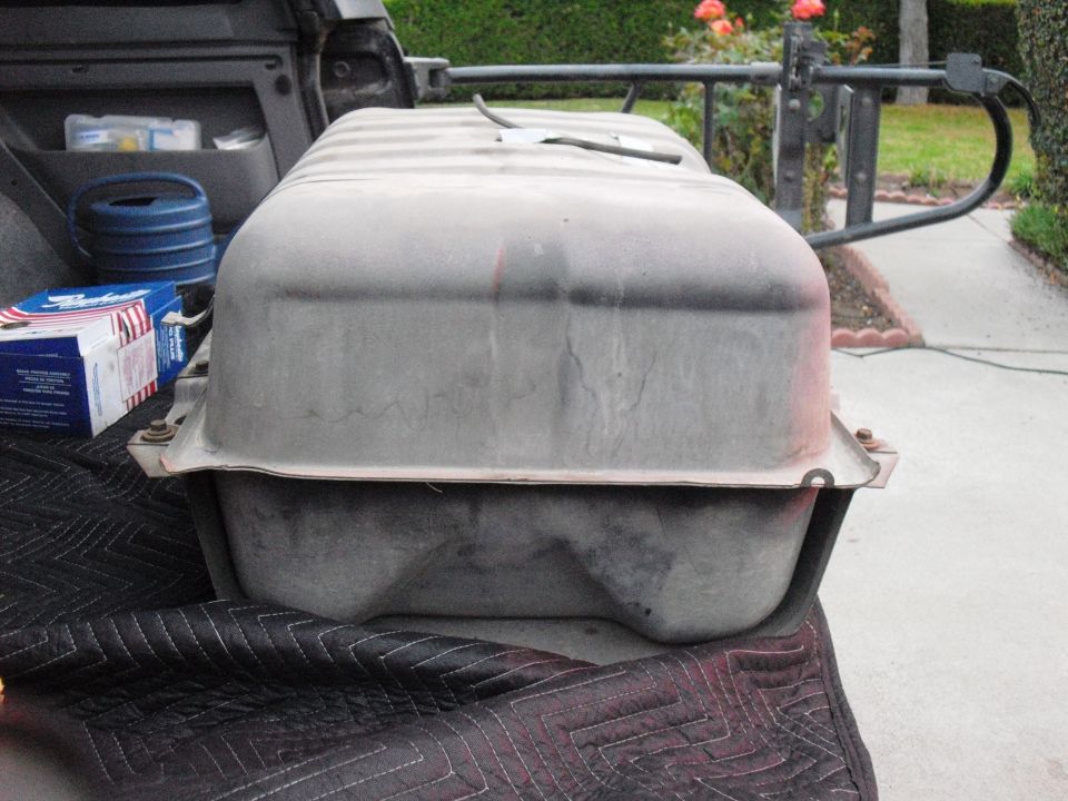 Gray metal fuel tank with ribbed top, resting on a black quilted mat in the bed of a pickup truck