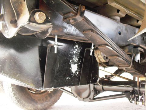 1986 Ford Bronco - Close underbody view of a fabricated black steel skid plate or tank shield mounted on threaded studs under the frame, with a multi-leaf spr…