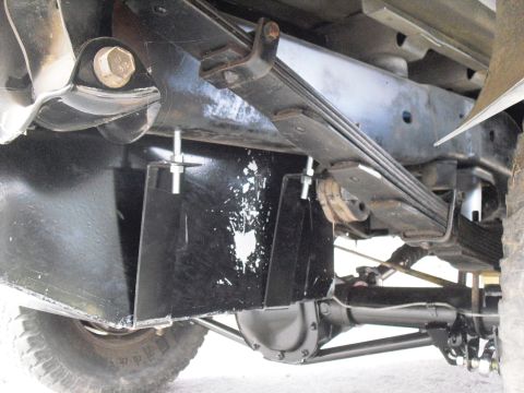 1986 Ford Bronco - Underbody shot looking up at a truck chassis, showing a fabricated black sheet-metal skid plate or fuel tank shield hung on threaded studs…