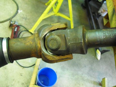 1986 Ford Bronco - A close-up view of a worn driveshaft universal joint, with a blue bucket and yellow equipment in the background.