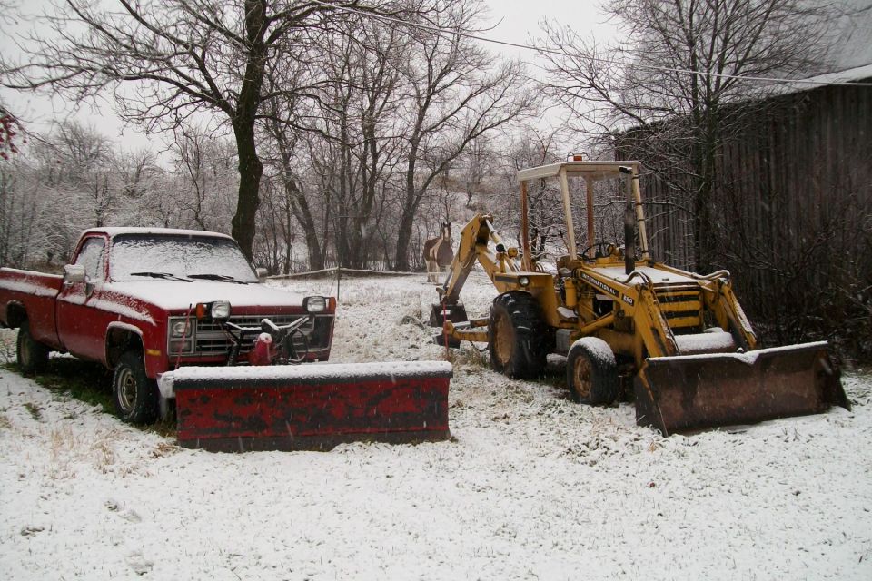 Red pickup with snow plow beside yellow backhoe loader in snowy yard