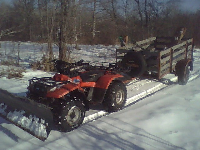 Orange ATV with front snow plow pulling a wood-rail utility trailer on snow