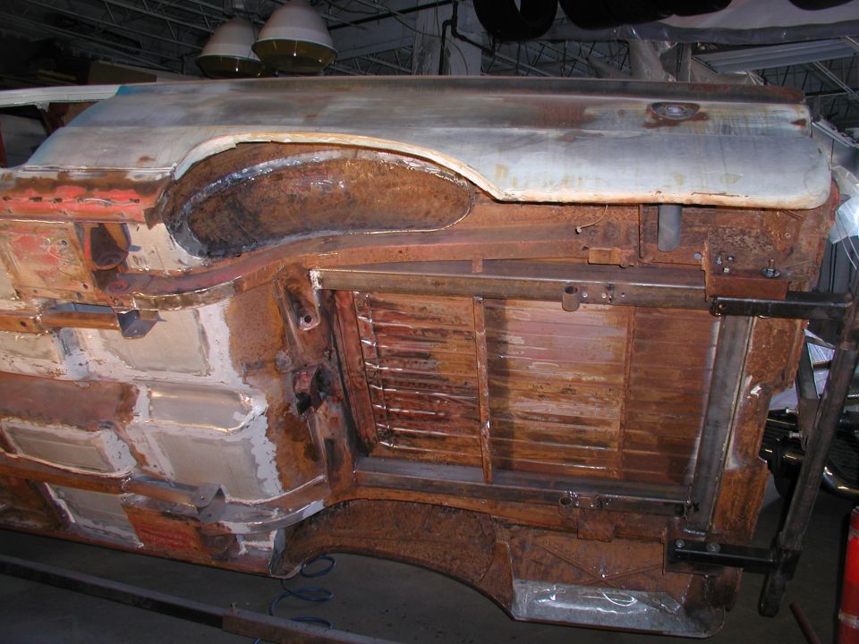 Vehicle body on a rotisserie showing rusty underside floor pans and primered quarter panel during restoration