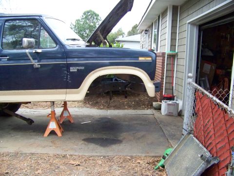 1985 Ford Bronco - A blue Ford Bronco is raised on jack stands in front of a garage, with its hood open.