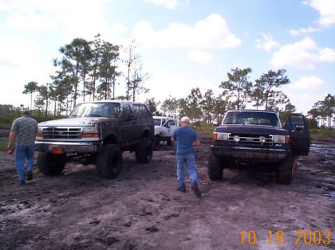 1995 Ford Bronco - Several off-road vehicles are parked in a muddy area with trees in the background under a cloudy sky.