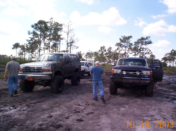 two lifted trucks covered in mud, one black Bronco and one dark pickup, with people standing around in a muddy field