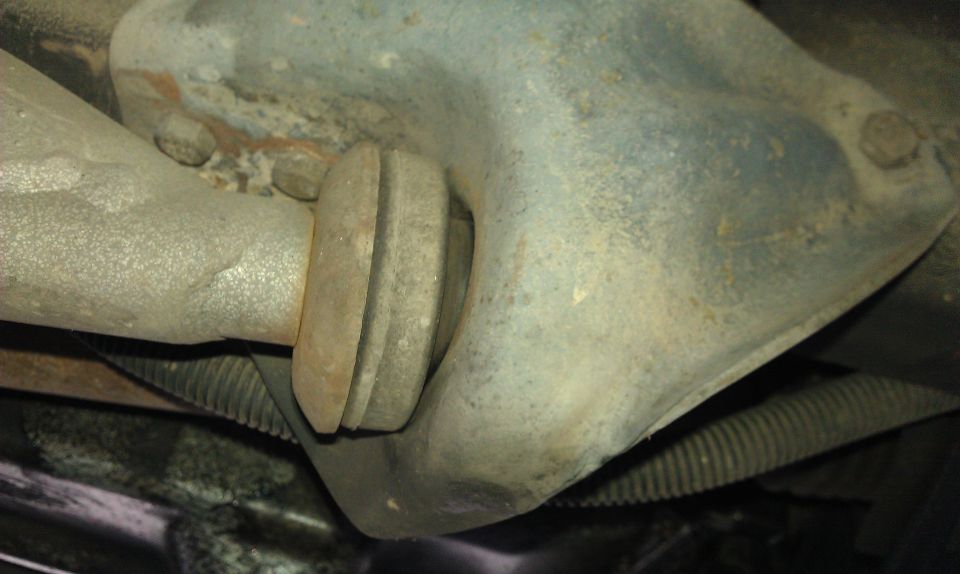 Underside close-up of a suspension arm with rubber bushing meeting a cast metal component, rusty and dirty.