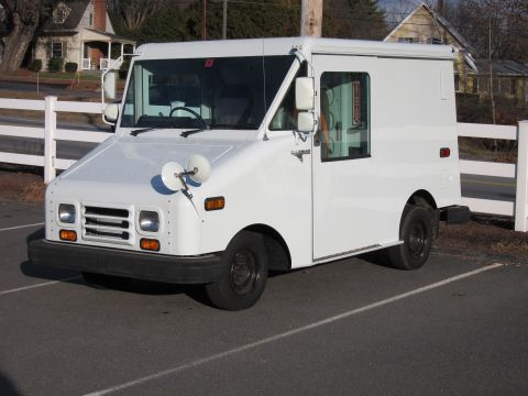 1991 Ford LTD Crown Victoria - A white Grumman LLV-style boxy mail/delivery van photographed from the front-left three-quarter angle, parked in a paved lot beside a white…