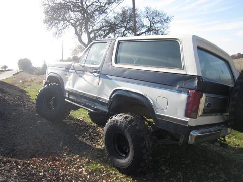 1985 Ford Bronco - This modified Ford Bronco features significant lift and oversized tires, parked on a dirt incline.