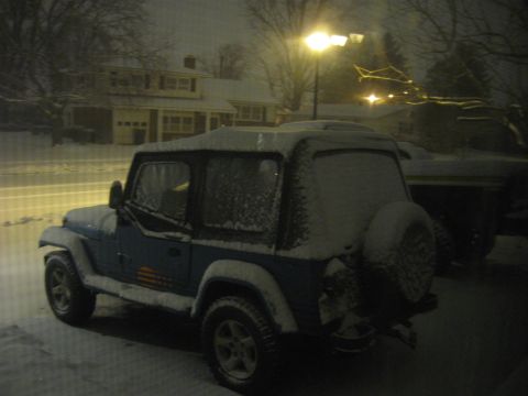 1991 Jeep YJ - A blue Jeep Wrangler is covered in snow, parked in front of a house during a snowfall at night.