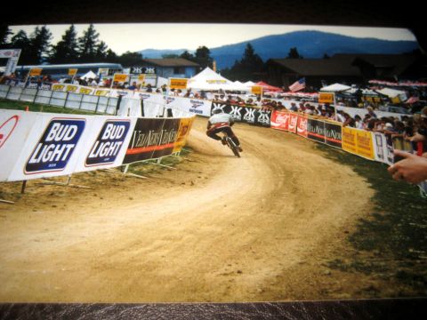 1996 Ford Bronco - A cyclist navigates a dirt track during a race, with spectators and sponsor banners lining the course.