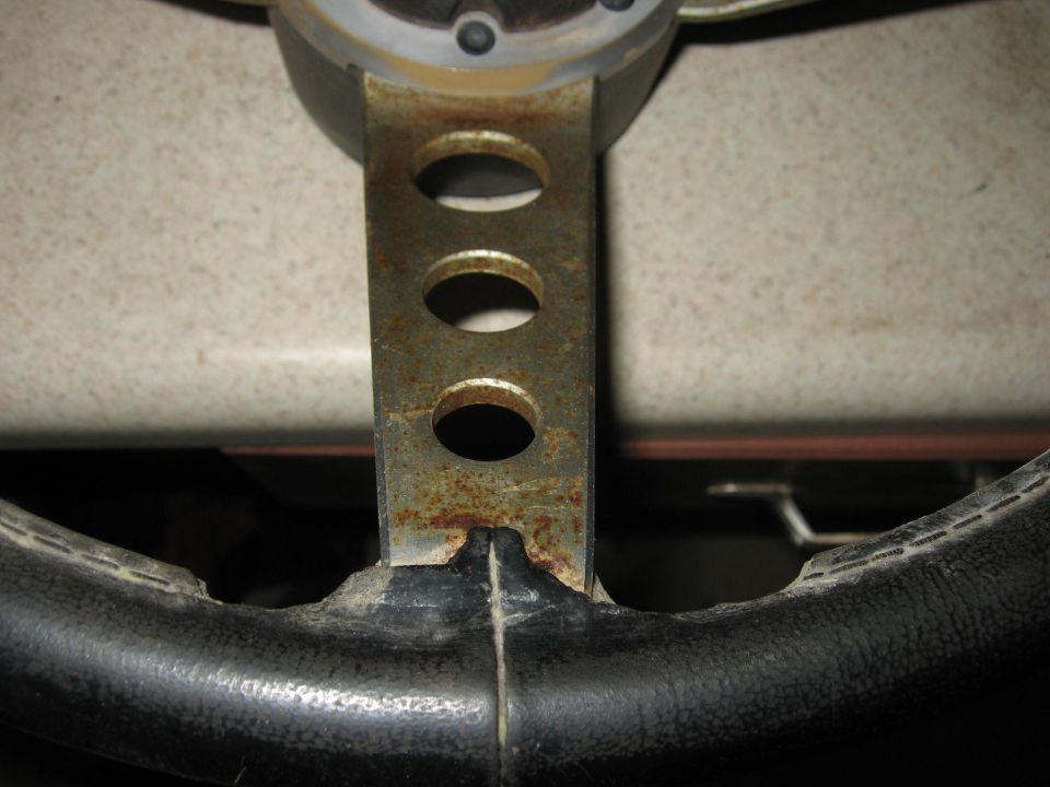 Close-up of a rusty metal steering wheel hub with three oval cutouts and black grip material