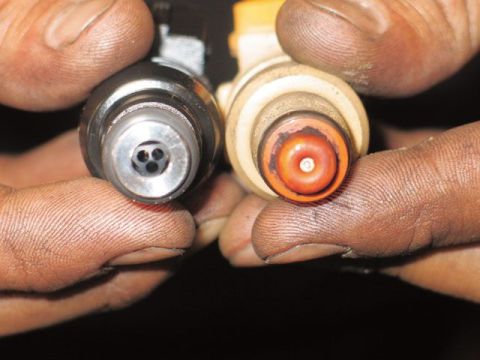 1990 Ford Bronco - Two fuel injectors held side by side, viewed from the nozzle end; the left one has a dark metal body with a multi-hole disc tip, while the…