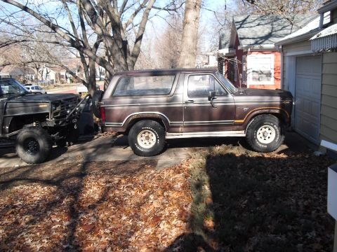 1986 Ford Bronco - A weathered dark brown/maroon 1986 Ford Bronco full-size SUV shown in side profile, parked on a leaf-covered driveway with chrome wheels an…