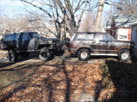 1986 Ford Bronco - Two full-size Ford Broncos parked side by side in a leaf-covered yard; the left one is a stripped project in black primer with a lifted sus…