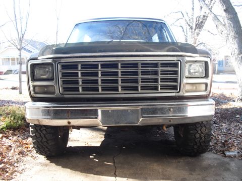1986 Ford Bronco - Head-on low view of a weathered brown 1980s full-size Ford truck front end on a cracked concrete driveway, showing the wide chrome-trimmed…