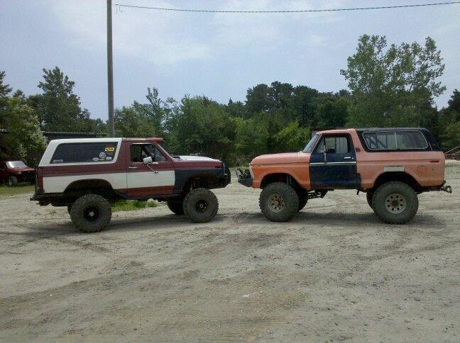 Two lifted Ford Broncos parked side-by-side, one red/white, one orange/blue