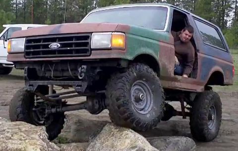 1988 Ford Bronco - A weathered green and rust-brown Ford Bronco full-size SUV with a lifted suspension and oversized mud tires crawls over large rocks, shown…