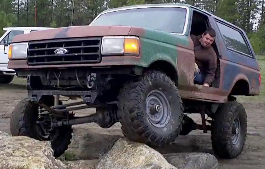 Lifted green/brown Ford Bronco on large mud tires crawling over rocks with driver leaning out.