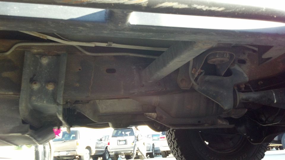 Underside view of a truck's frame and suspension components. Other vehicles are visible in the background