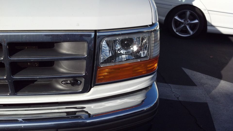 Close-up of white truck's front grille and headlight assembly. Another white car is visible in the background