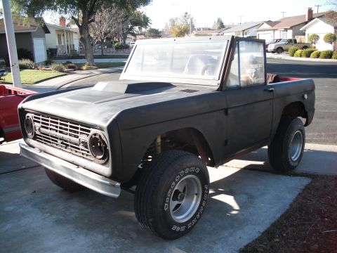 1968 Ford Bronco - Three-quarter front view of an early Ford Bronco in flat black primer on a suburban driveway, doorless and topless with the windshield fram…
