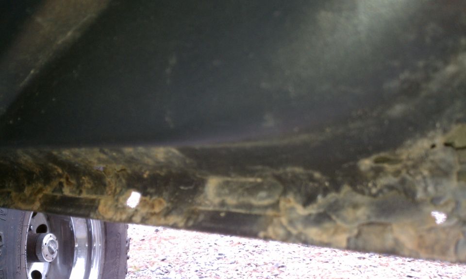 Undercarriage of a vehicle showing significant rust and dirt, with a tire visible on the left