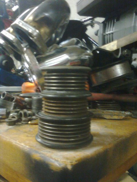 1984 Ford Bronco - A grooved multi-ribbed pulley or stacked pulley assembly sits on a wooden workbench surface, with various metal automotive parts and clamps…