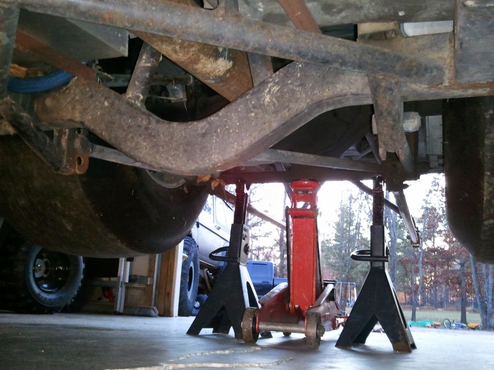Underside of a rusty framed vehicle on jack stands with a red floor jack