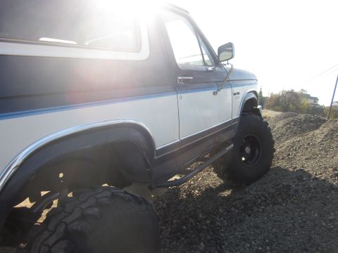 1985 Ford Bronco - A lifted 1985 Ford Bronco is shown from a three-quarter front angle on a gravelly surface.
