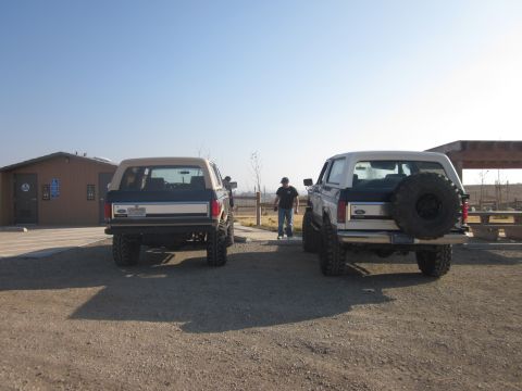 1985 Ford Bronco - Two Ford Broncos are parked in a lot near a building and picnic area under a clear sky.