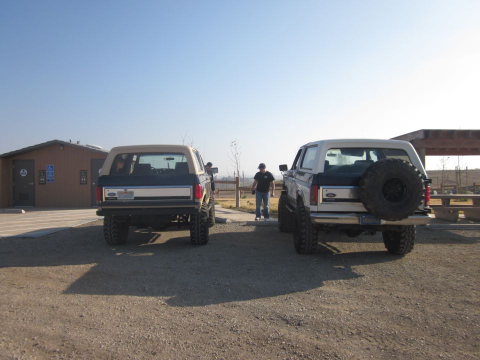 Two Ford Broncos parked rear-view, one with a spare tire mounted on the back. A man stands between them