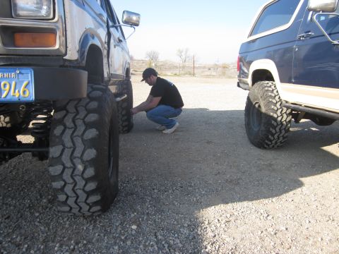 1985 Ford Bronco - Two lifted Ford Broncos are parked side-by-side on a gravel lot, with a man attending to one of the tires.