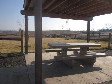 1985 Ford Bronco - A concrete picnic table sits under a wooden shelter in a park-like setting with a fence and dry grass visible.