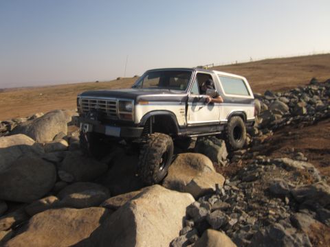 1985 Ford Bronco - A modified Ford Bronco drives over a rocky path under a clear sky.