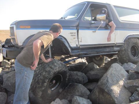1985 Ford Bronco - A man inspects the large tire of a 1985 Ford Bronco as it maneuvers over a boulder-strewn path.