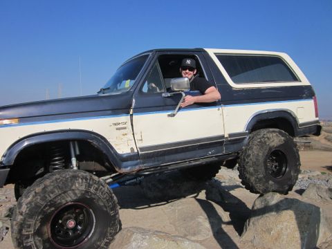 1985 Ford Bronco - A man drives a modified Ford Bronco over rocks under a clear blue sky.