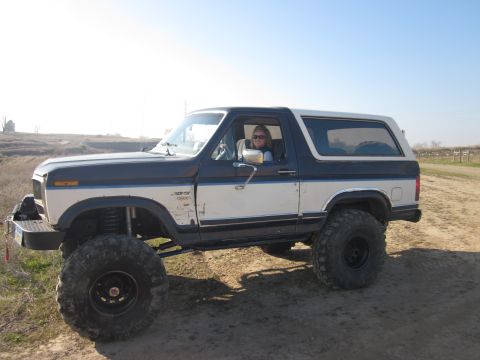 1985 Ford Bronco - A woman drives a lifted two-tone blue and white Ford Bronco off-road.