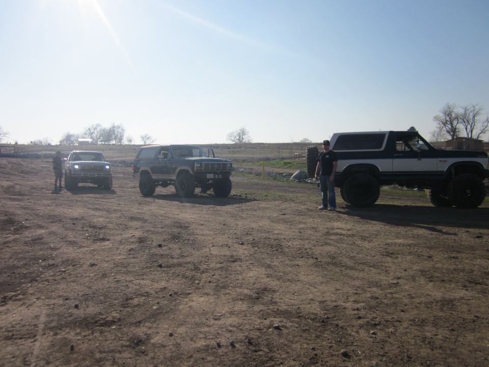 Three lifted SUVs parked in a dirt lot under a bright sky. The Bronco on the right is black and white with large tires