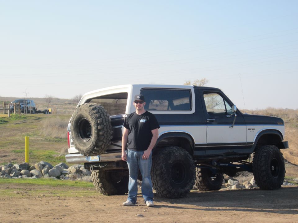 Man standing next to a lifted 1985 Ford Bronco with a spare tire mounted on the rear. The Bronco is white and dark blue