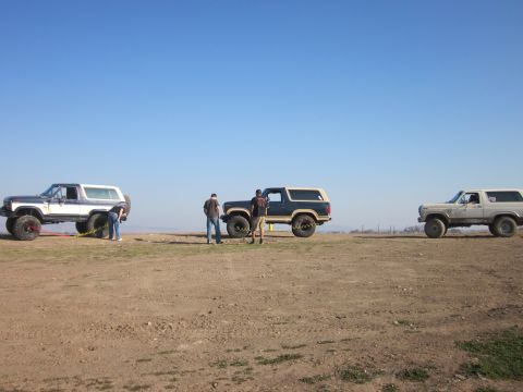 1985 Ford Bronco - Three Ford Broncos are parked on a dirt lot with people standing around them.