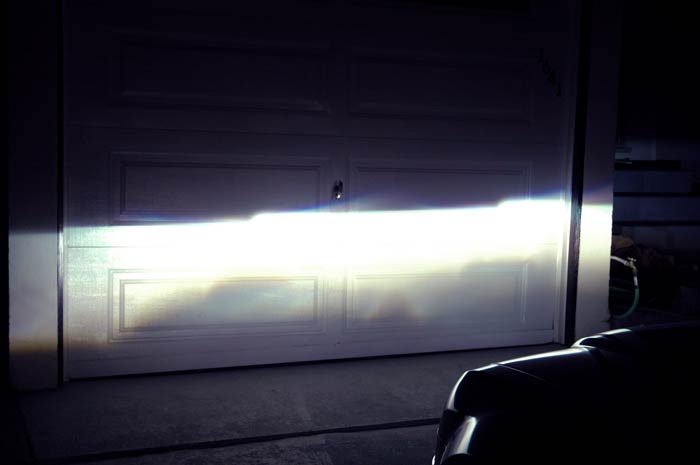 Headlights illuminate a garage door, with a portion of a dark vehicle in the foreground