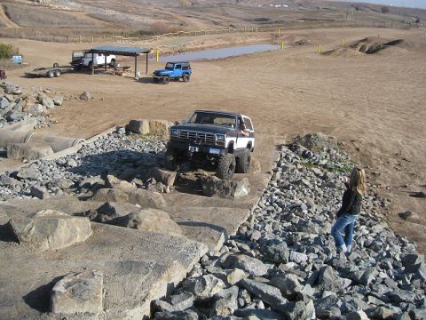 1985 Ford Bronco - A modified Ford Bronco navigates a rocky obstacle course, with other vehicles and a pond in the distance.