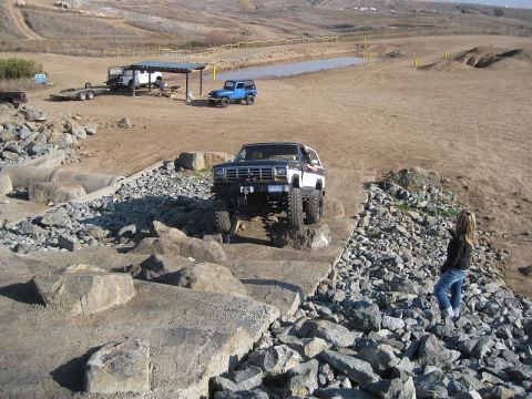 1985 Ford Bronco - A modified Ford Bronco navigates a challenging off-road course with a woman observing.