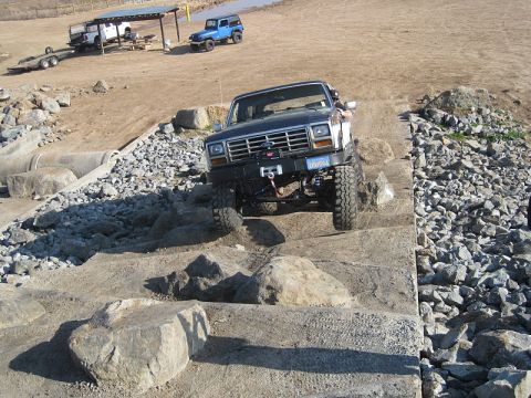 1985 Ford Bronco - A dark blue Ford Bronco navigates a rocky obstacle course, with other vehicles visible in the background.