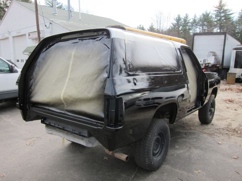 1987 Dodge 1500 - This black SUV is covered in plastic sheeting and tape, likely for a paint job, parked outdoors.