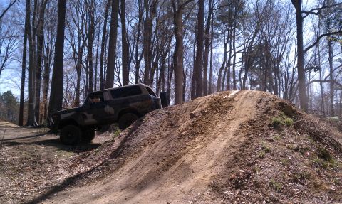 1979 Ford Bronco - A camouflaged SUV navigates a dirt track in a forest setting.