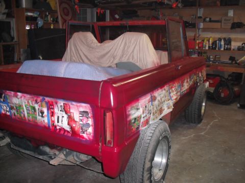 1968 Ford Bronco - A red SUV is parked in a garage, its side panels covered with taped-on images, possibly for a custom paint job.