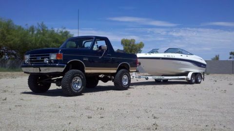 1991 Ford Bronco - A lifted dark blue Ford Bronco tows a white boat on a trailer under a clear blue sky.