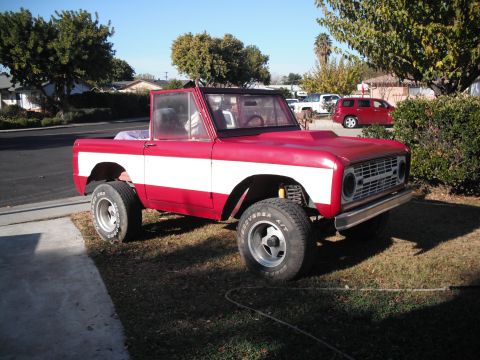 1968 Ford Bronco - A red and white 1968 Ford Bronco is parked on a grassy area next to a street, with other vehicles visible in the background.